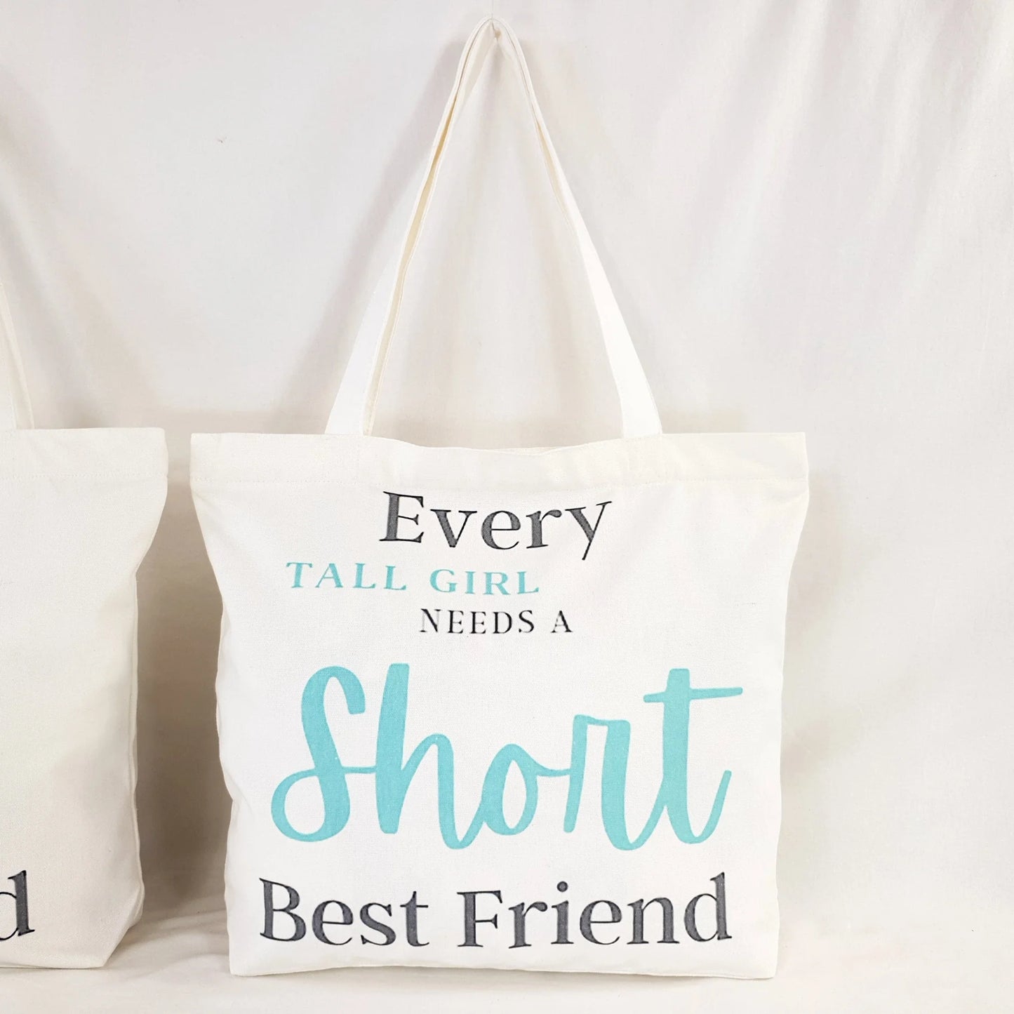 Tall Friend and Short Friend bags, one bag print text reads : "Every Short girl needs a tall best friend" and the words "short girl" and "Tall" is in Teal color and rest of writing is in dark grey. the other bag print reads: "Every tall girl needs a short best friend" colouring of the print text is the same as the other bag.  Bag itself is white with shoulder straps, zipper on the top