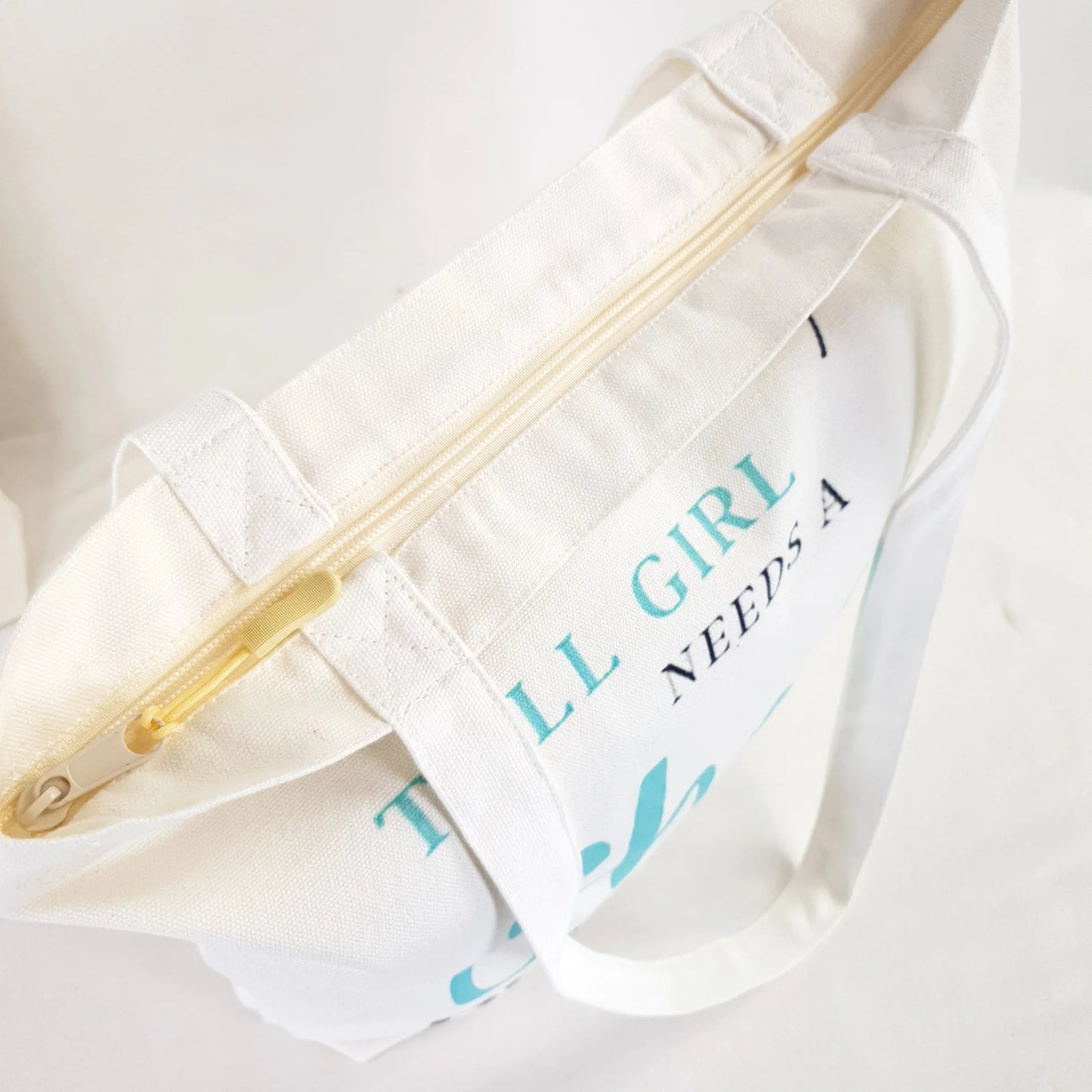 Tall Friend and Short Friend bags, one bag print text reads : "Every Short girl needs a tall best friend" and the words "short girl" and "Tall" is in Teal color and rest of writing is in dark grey. the other bag print reads: "Every tall girl needs a short best friend" colouring of the print text is the same as the other bag.  Bag itself is white with shoulder straps, zipper on the top